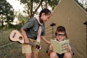 Kids in scout uniforms enjoying a camping experience with a guitar and scout field book outdoors.